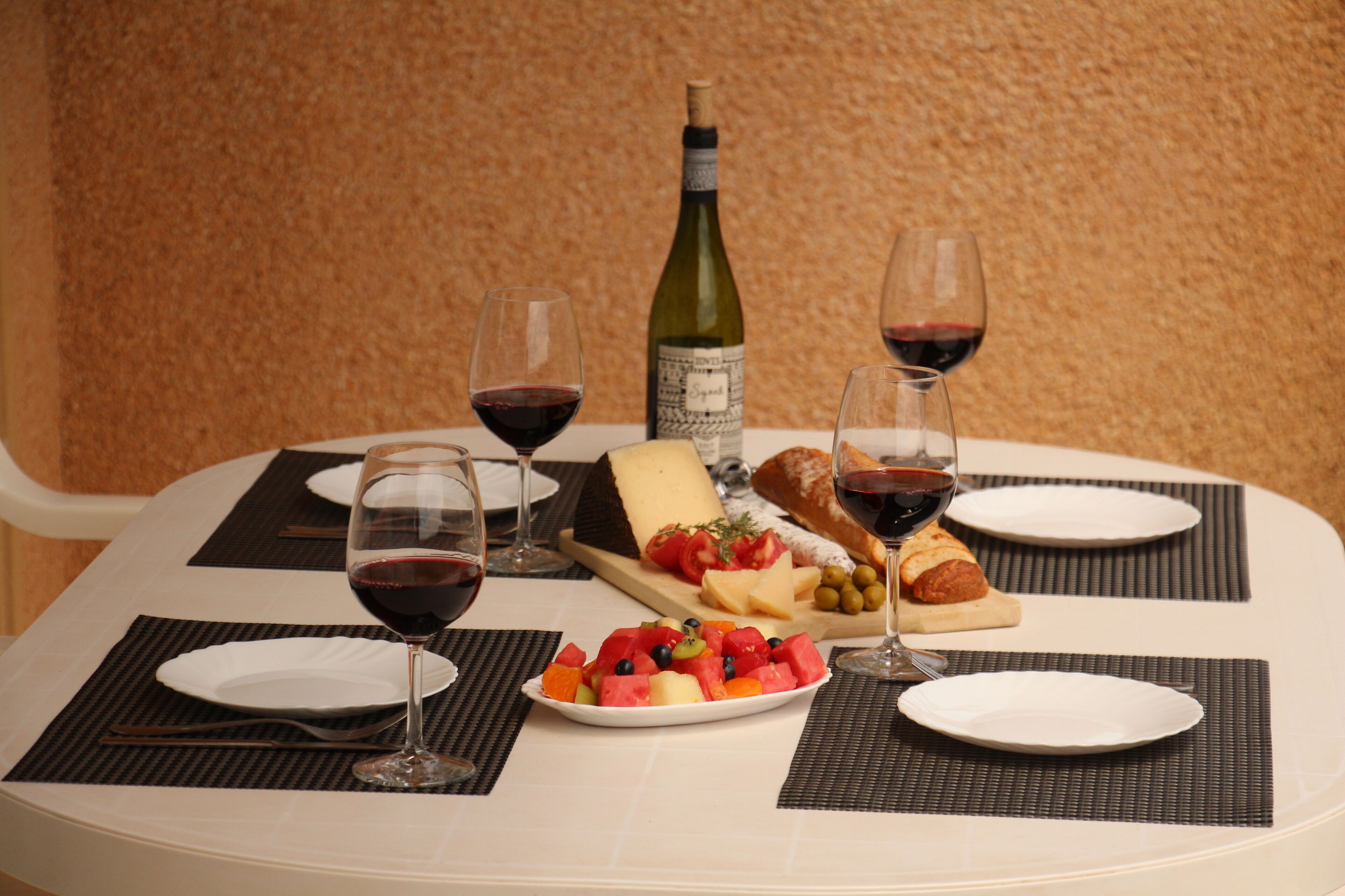 A dining table set with plates, cutlery, a bottle of wine, glasses, a cheese board, and a fruit platter.