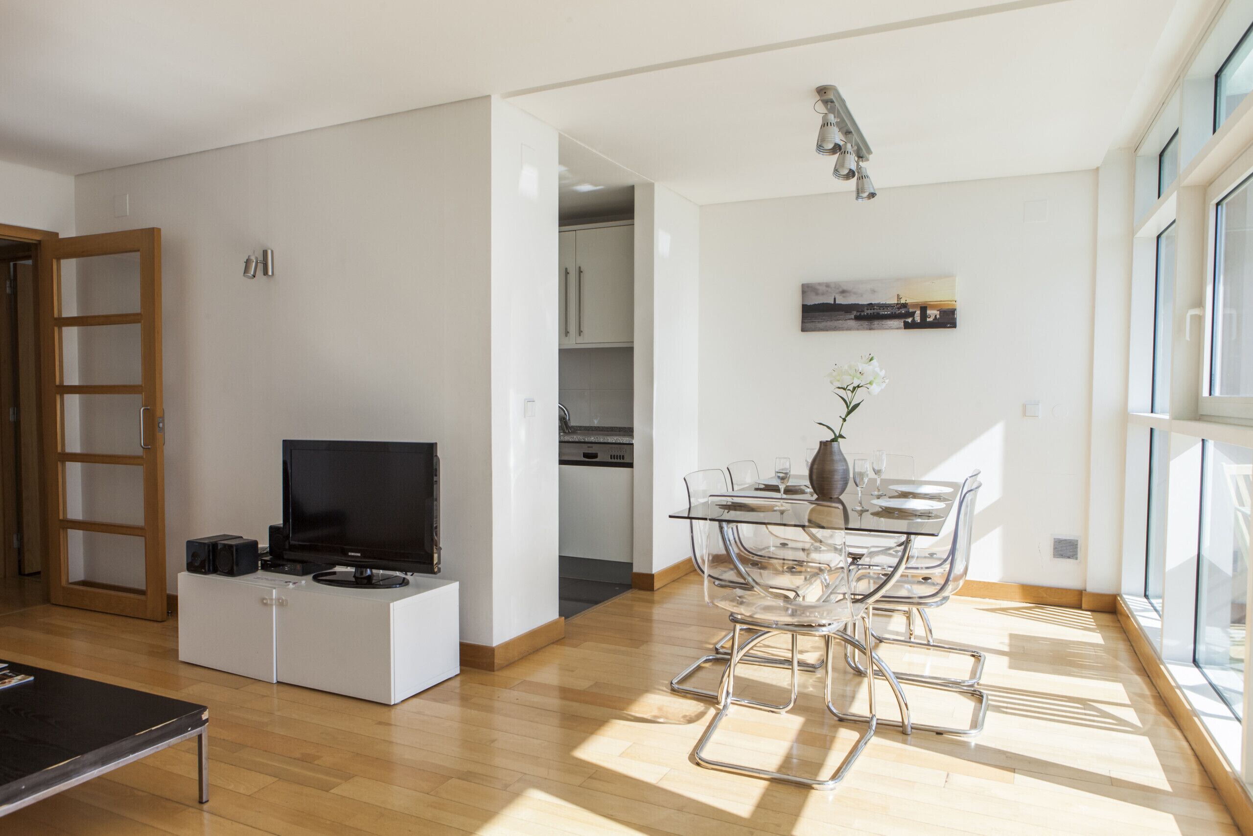 A modern living room with a flat-screen TV, a glass dining table, and a kitchen in the background.