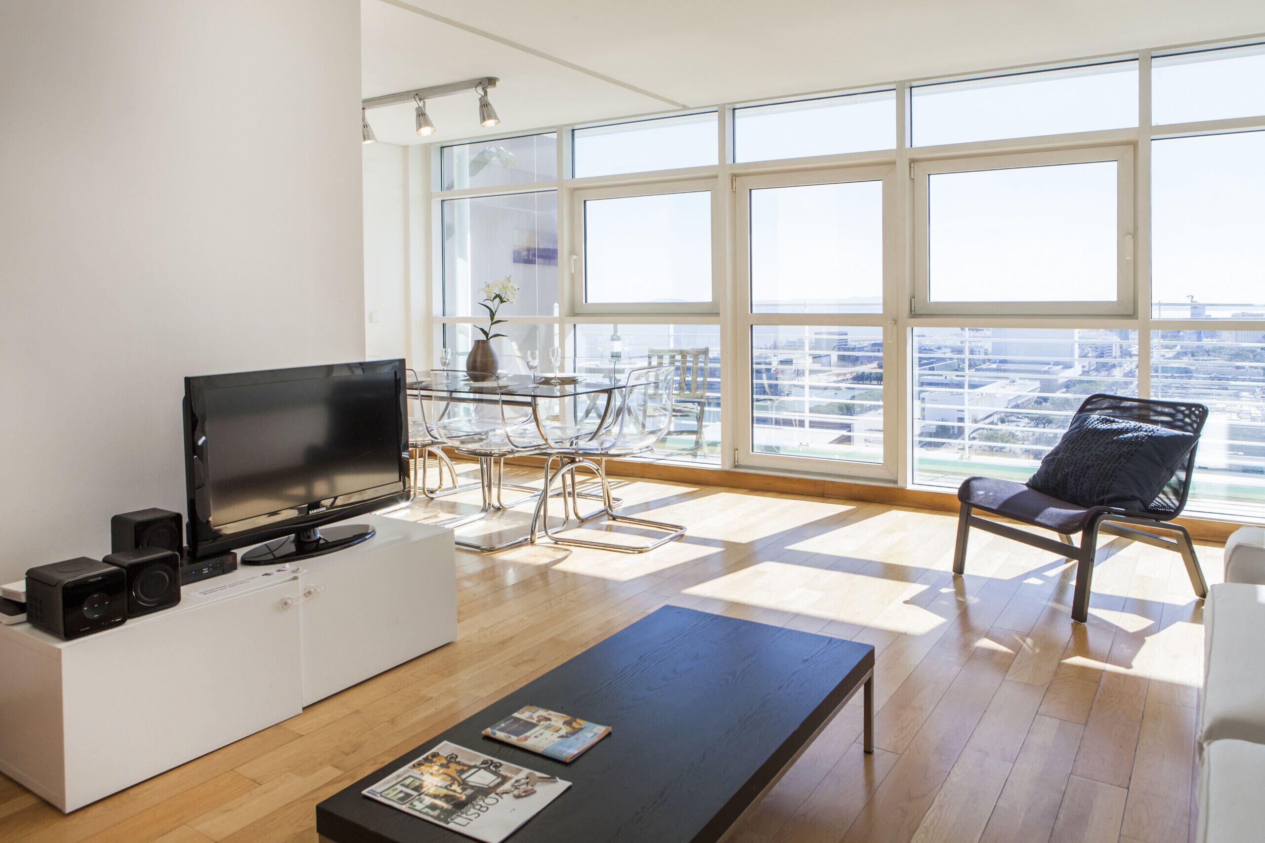 Een moderne woonkamer met een flatscreen-televisie, een salontafel met tijdschriften, een eethoek met een glazen tafel en stoelen, en grote ramen met uitzicht over de stad.