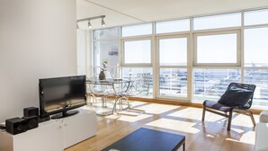 A modern living room with a flat-screen TV, a coffee table with magazines, a dining area with a glass table and chairs, and large windows offering a city view. - RENT4REST LISBON PARQUE DAS NAÇÕES 17TH FLOOR RIVER VIEW (Lisboa ciudad)
