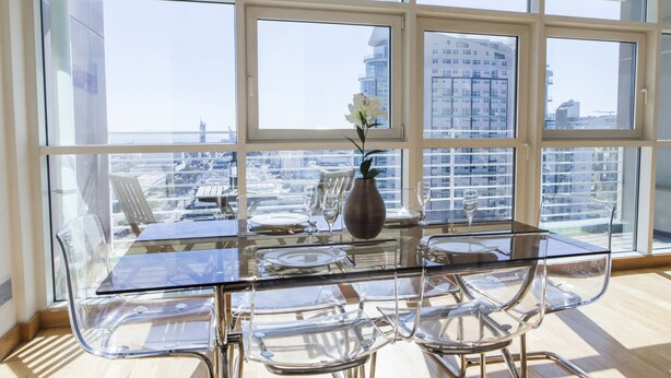 A dining area with a glass table and chairs, a vase with flowers, and a view of a cityscape through large windows. - RENT4REST LISBON PARQUE DAS NAÇÕES 17TH FLOOR RIVER VIEW (Lisboa ciudad)
