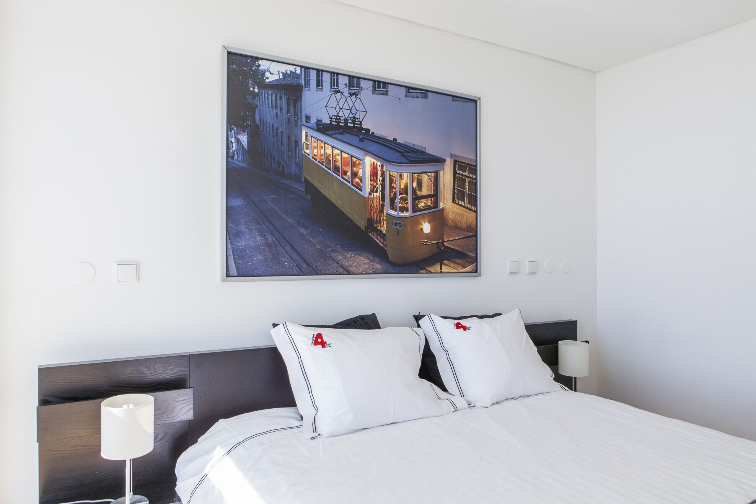 A hotel room with a large framed picture of a tram, a neatly made bed with white bedding, and a modern bedside table with a lamp.