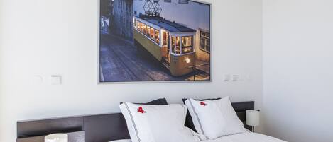 A hotel room with a large framed picture of a tram, a neatly made bed with white bedding, and a modern bedside table with a lamp.