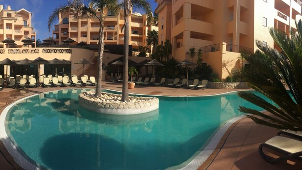 A pool area with lounge chairs and umbrellas, surrounded by palm trees and a multi-story building with balconies. - Estrela da Luz Spectacular Penthouse With Sea And Pool View (Luz)