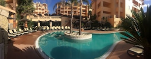 Un espace piscine doté de chaises longues et de parasols, entouré de palmiers et bordé d’un immeuble à plusieurs étages avec des balcons.