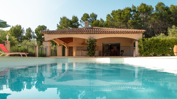 A swimming pool with a covered patio area, a red lounge chair, and a view of trees in the background. - House / Villa - DRAGUIGNAN (Draguignan)