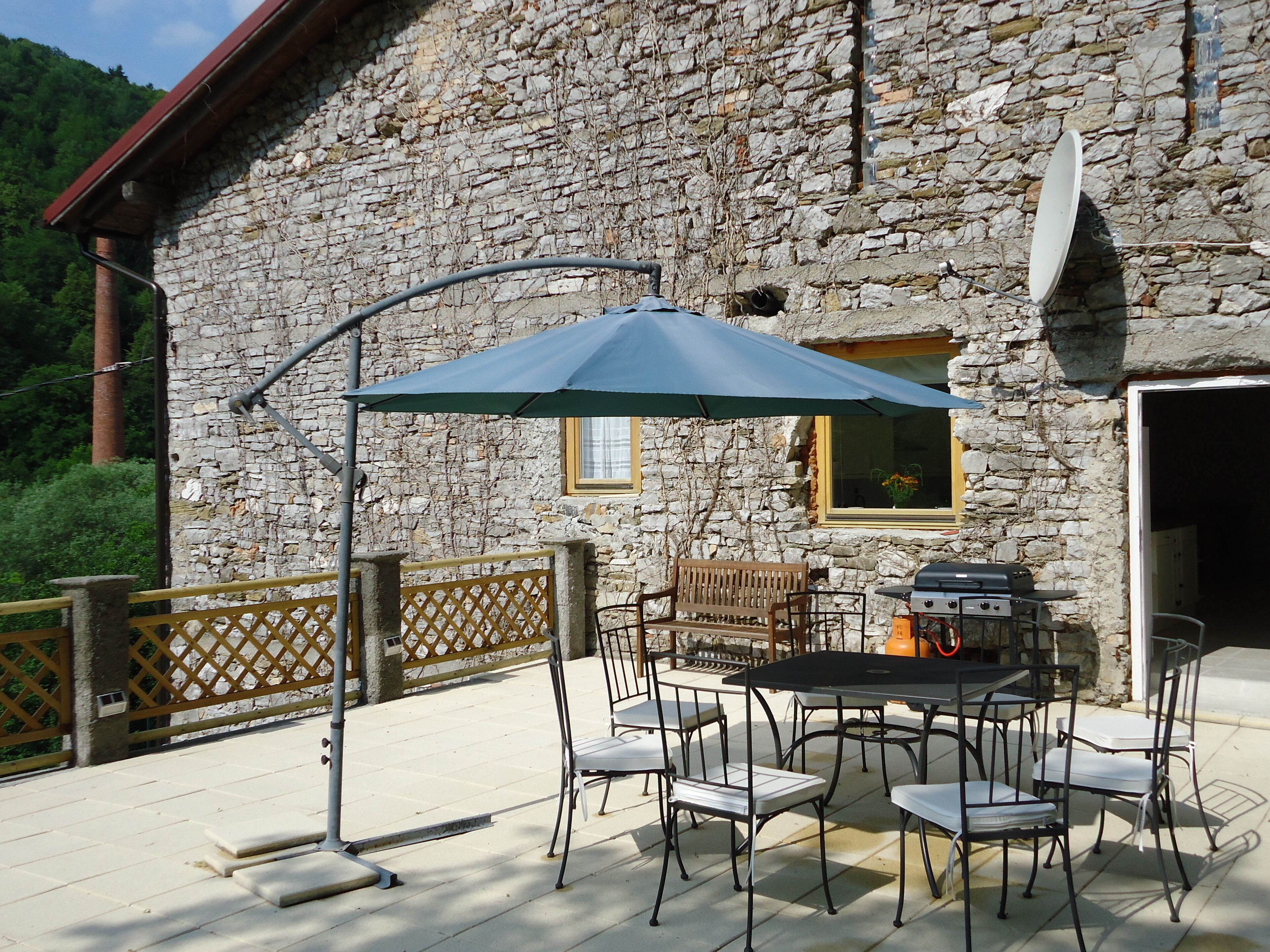 An outdoor patio with a stone building, a table with chairs, a grill, and a blue umbrella.