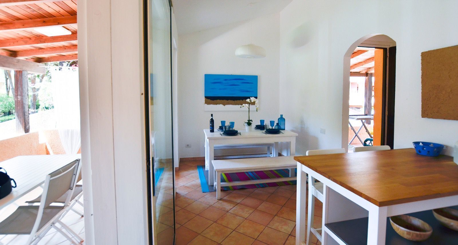 A bright room with a white table, a wooden table, and a bench. There is a blue painting on the wall.