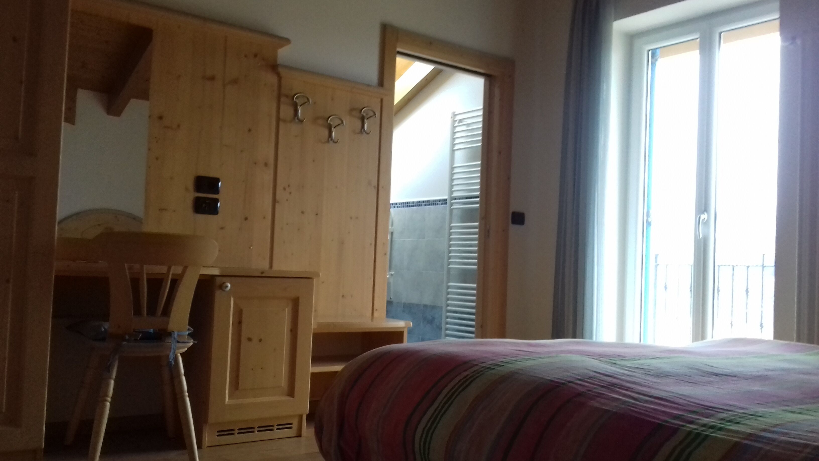 A bedroom with a wooden wardrobe, a bed with a striped blanket, a chair, and a window with a view of the outside.