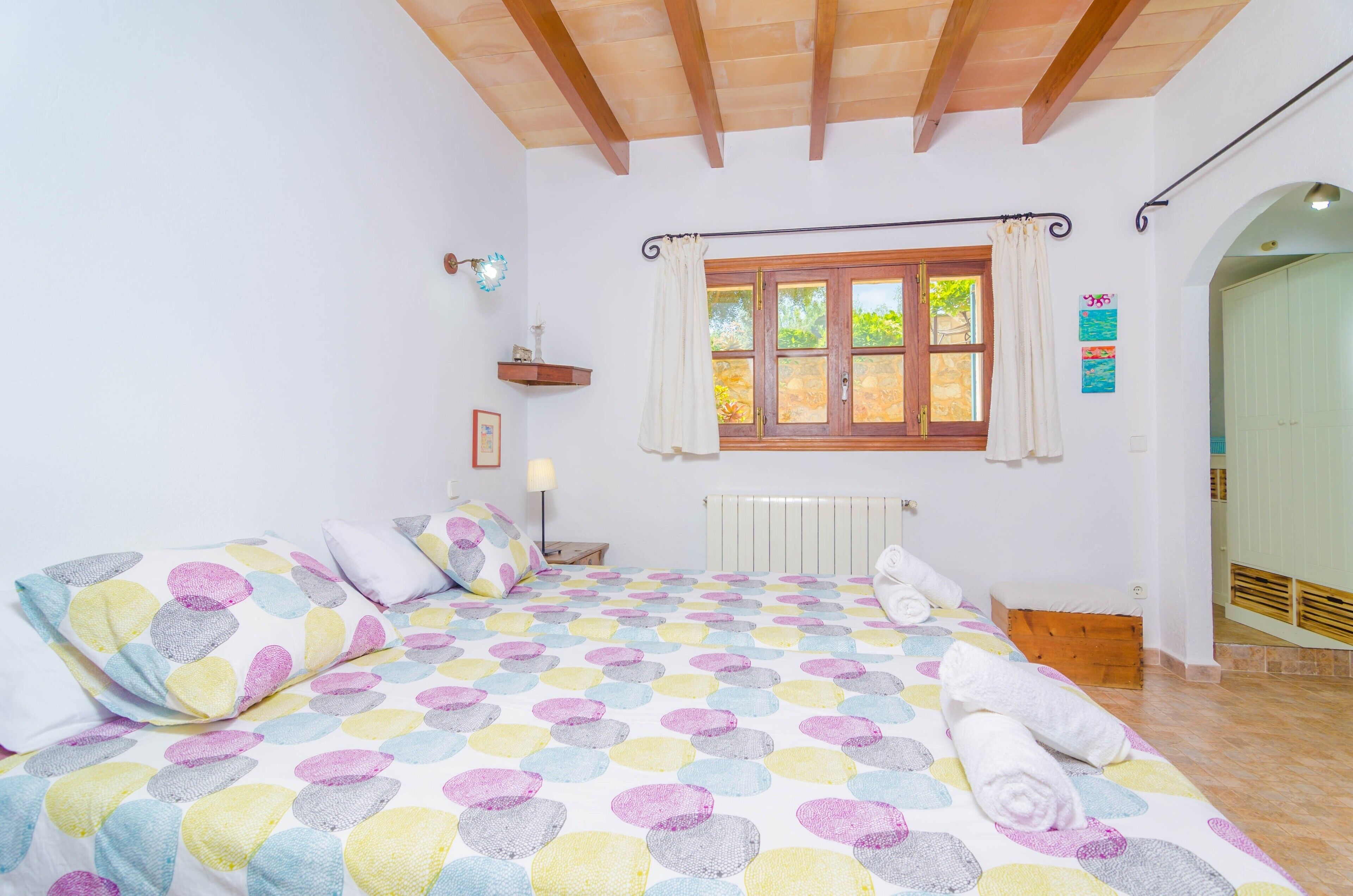 Een slaapkamer met een bed, een raam met gordijnen, een radiator, een houten bankje en een plank met een lamp.