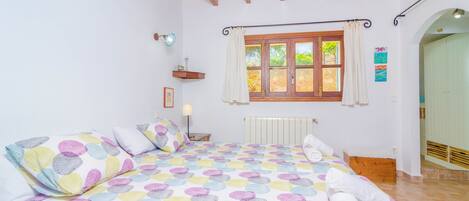 Een slaapkamer met een bed, een raam met gordijnen, een radiator, een houten bankje en een plank met een lamp.