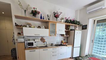 A modern kitchen with white cabinets, a microwave, and a refrigerator. There are shelves with decorative items and a dining table with chairs.