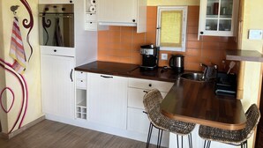 A compact kitchen with white cabinets, a coffee maker, a kettle, and a small dining area with wicker chairs.
