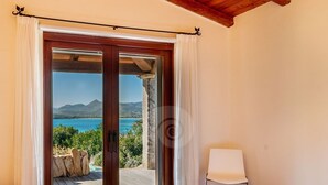 Ein Schlafzimmer mit Holzboden, einem Bett, einem weißen Stuhl und einer Glastür, die auf einen Balkon mit Blick auf das Meer und die Berge führt.