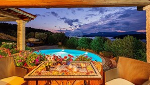 Ein Poolbereich mit einem Mosaik-Tisch, Korbmöbeln und Blick auf Berge und einen See im Abendlicht.