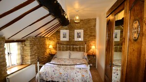 A bedroom with a stone wall, a wooden ceiling, a bed with a floral bedspread, two wall-mounted lamps, and a window with curtains.