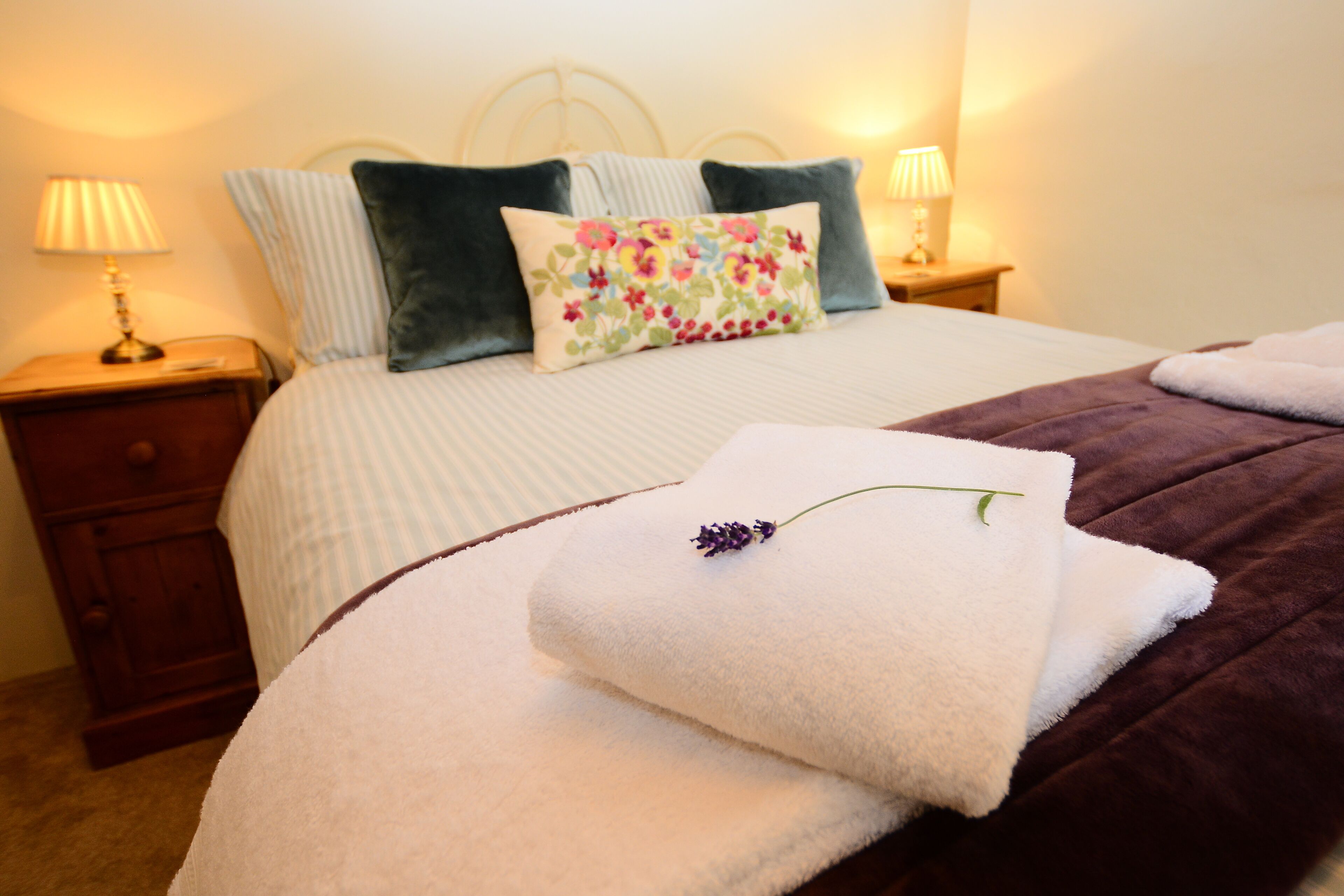 Una cama bien hecha con un cojín floral, una manta morada y una toalla blanca con una flor de lavanda. Hay dos lámparas de noche y una mesita de noche de madera con una lámpara encima.