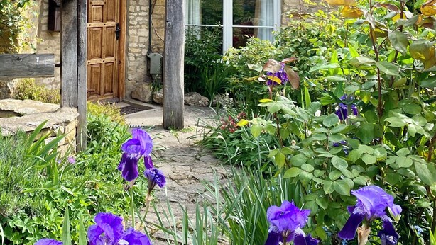 A stone house with a wooden door, a window, and a garden with purple flowers.