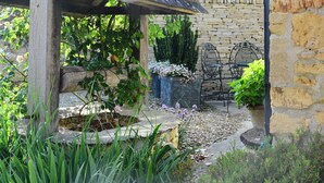A rustic garden with a stone wall, wooden pergola, and a seating area with metal chairs.
