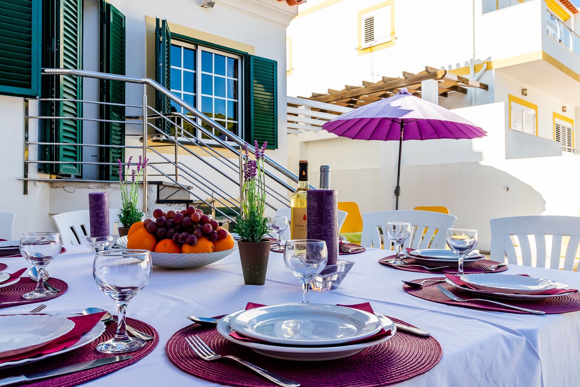 An outdoor dining area with a table set for two, featuring a white tablecloth, red placemats, and a purple umbrella.