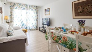 Ein helles Wohnzimmer mit einer Couch, einem Esstisch und Stühlen. Ein Fernseher steht auf einem Schrank, ein Fenster mit floralem Vorhang und ein gerahmtes Bild sind an der Wand zu sehen.