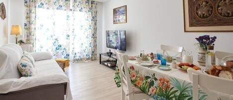 A well-lit living room with a sofa, dining table, and chairs. There is a TV on a stand, a window with floral curtains, and a framed picture on the wall.