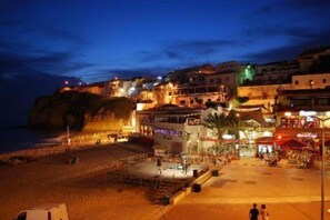 Una cittadina costiera di notte, con edifici lungo la scogliera, una spiaggia in primo piano e alcune persone che passeggiano.