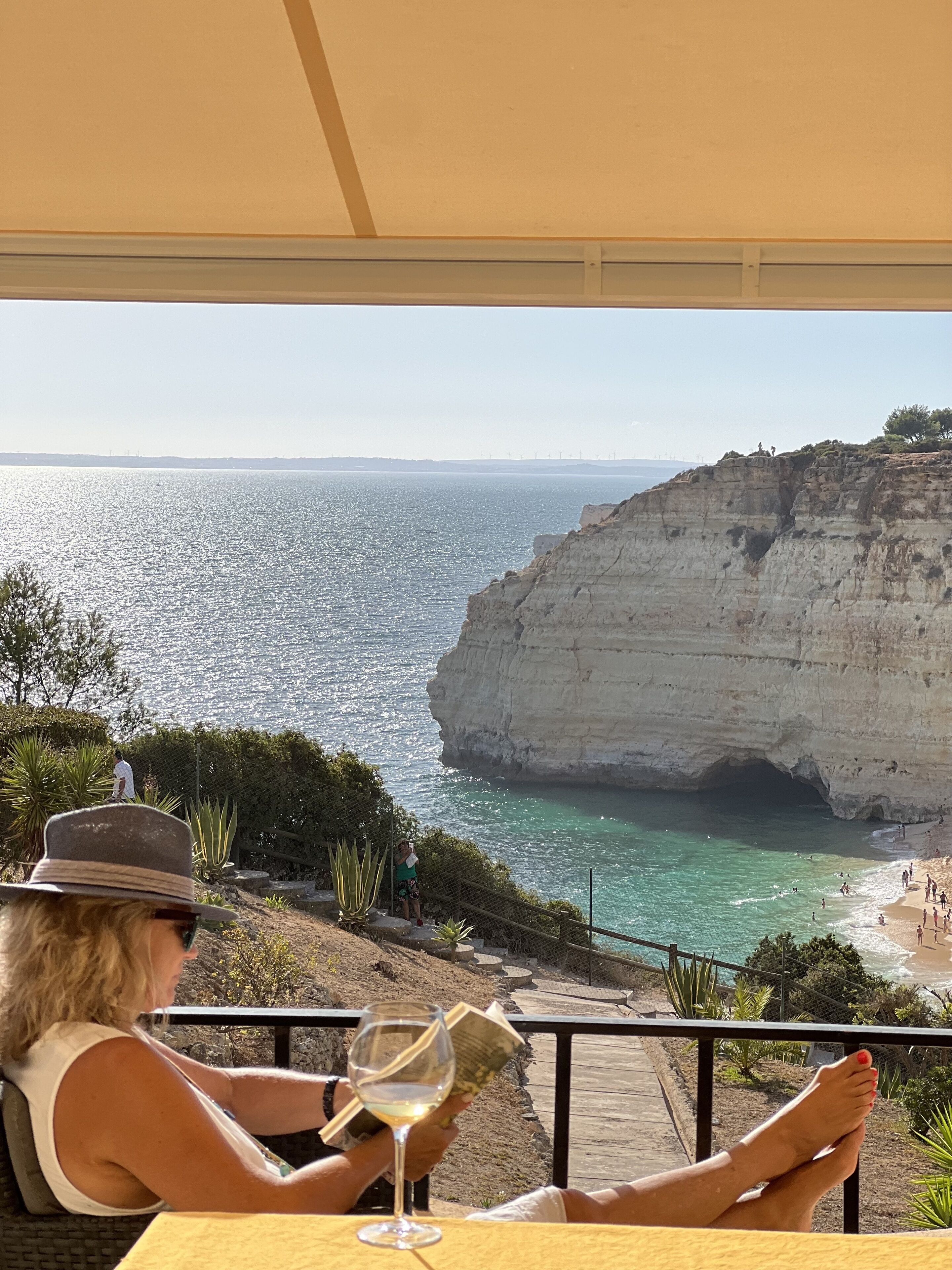 A person seated at a table with a glass of white wine, overlooking a coastal cliff and beach.