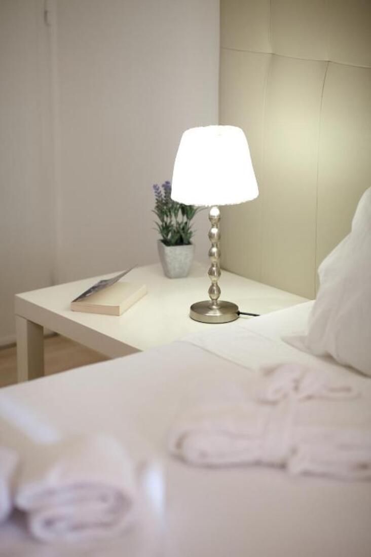 Una cama bien hecha con una almohada blanca y una toalla doblada. Una mesita de noche sostiene una lámpara, un libro y una pequeña planta en maceta.