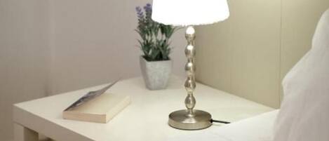 A neatly made bed with a white pillow and a folded towel. A bedside table holds a lamp, a book, and a small potted plant.