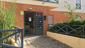 Entrada de um edifício com portão de metal, parede de tijolos e área pavimentada.