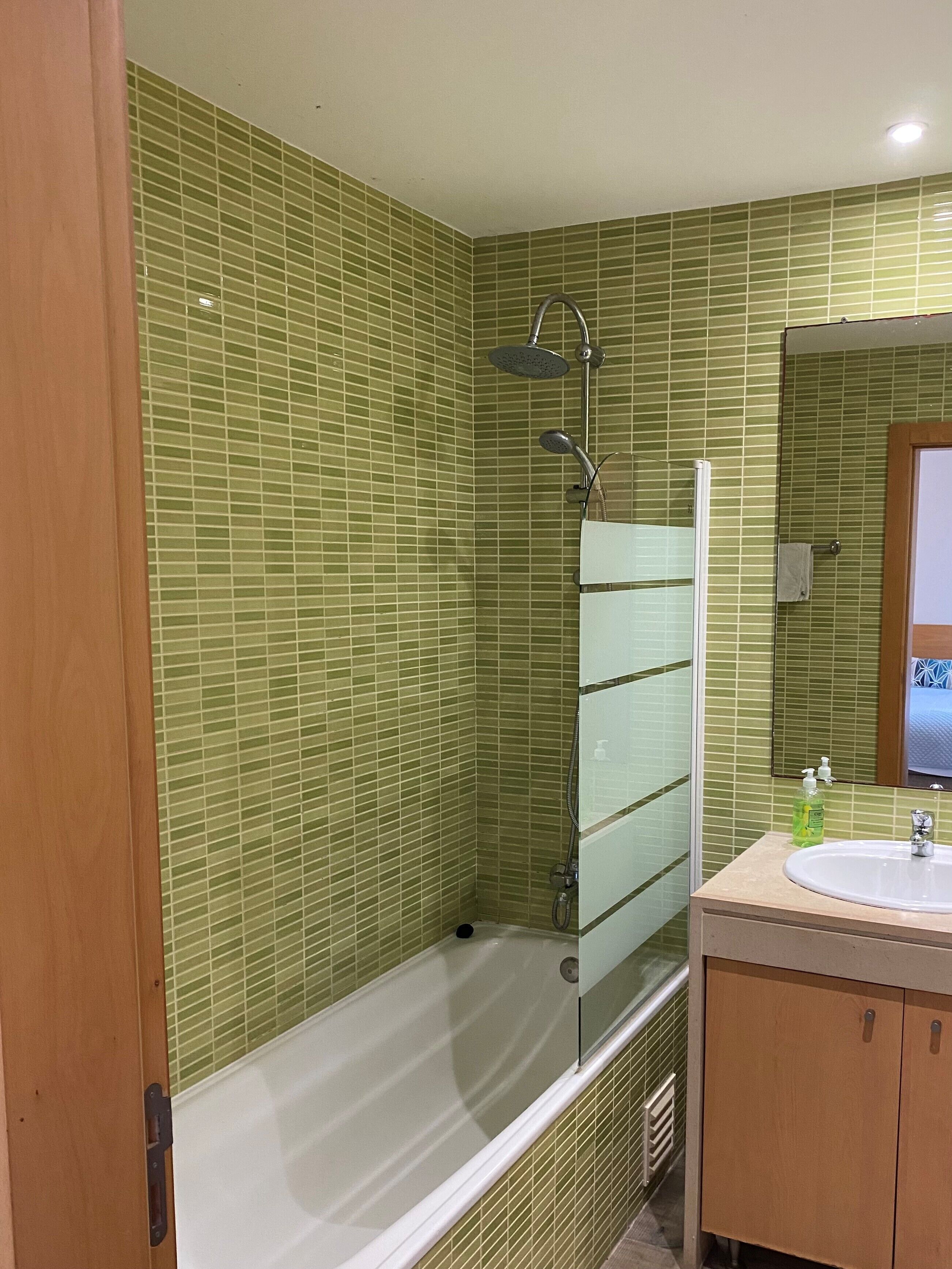 A bathroom with green tiled walls, a glass shower enclosure, a white bathtub, and a wooden vanity with a sink.