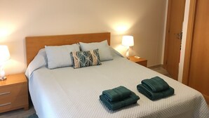 Cama bem arrumada com um edredom branco, dois travesseiros e duas toalhas dobradas sobre a cama.