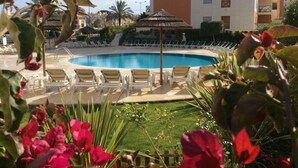 Piscina com espreguiçadeiras e guarda-sóis, cercada por vegetação e um edifício com varandas.