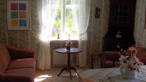 A room with floral wallpaper, a window with lace curtains, a framed picture, a small table with a vase of flowers, and two sofas.