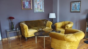 Un salon avec un canapé jaune, une petite table ronde et une table basse en verre. Des tableaux encadrés ornent le mur, et un vase rempli de fleurs rouges est posé sur une table d’appoint.