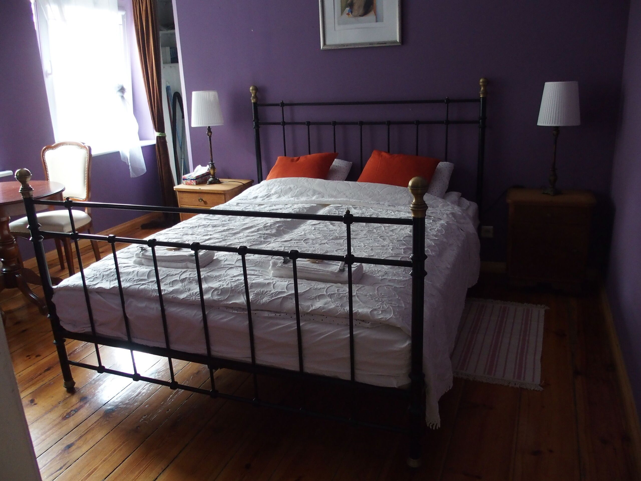 A bedroom with a metal bed frame, a wooden floor, a nightstand, a lamp, and a chair.