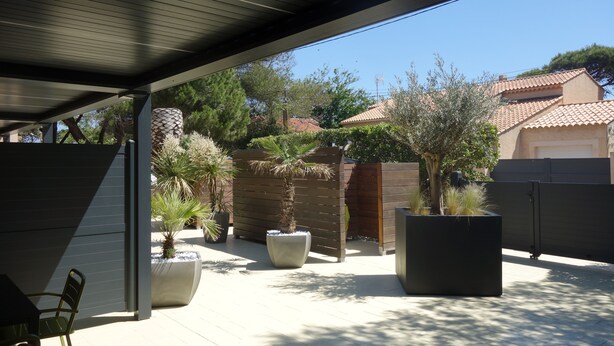Ein überdachter Patio-Bereich mit großen Pflanzkübeln, einem Holzzaun und Blick auf benachbarte Häuser.
