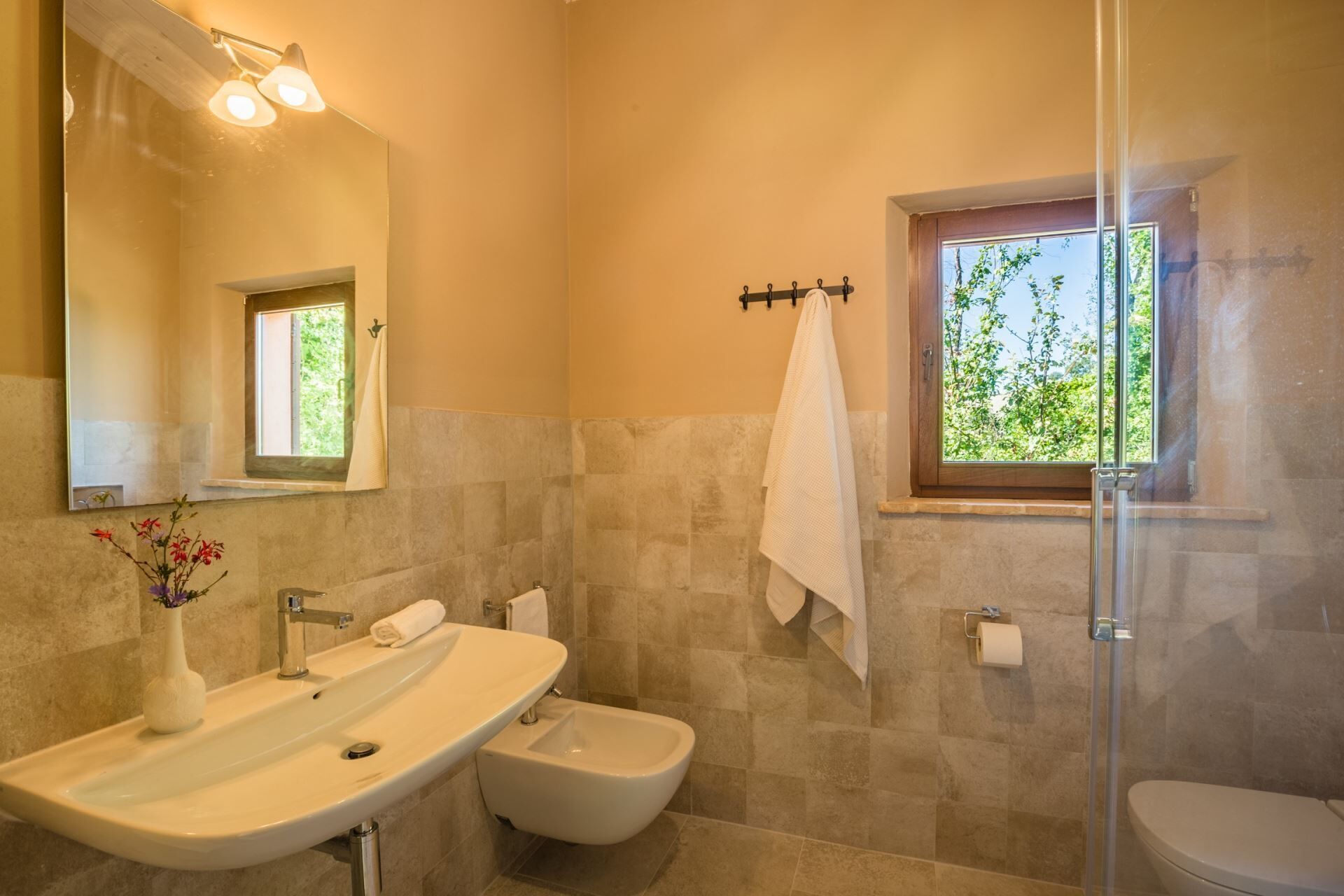 Ein Badezimmer mit Waschbecken, Bidet und Toilette, einem Spiegel und einem Fenster mit Blick auf Grünflächen.