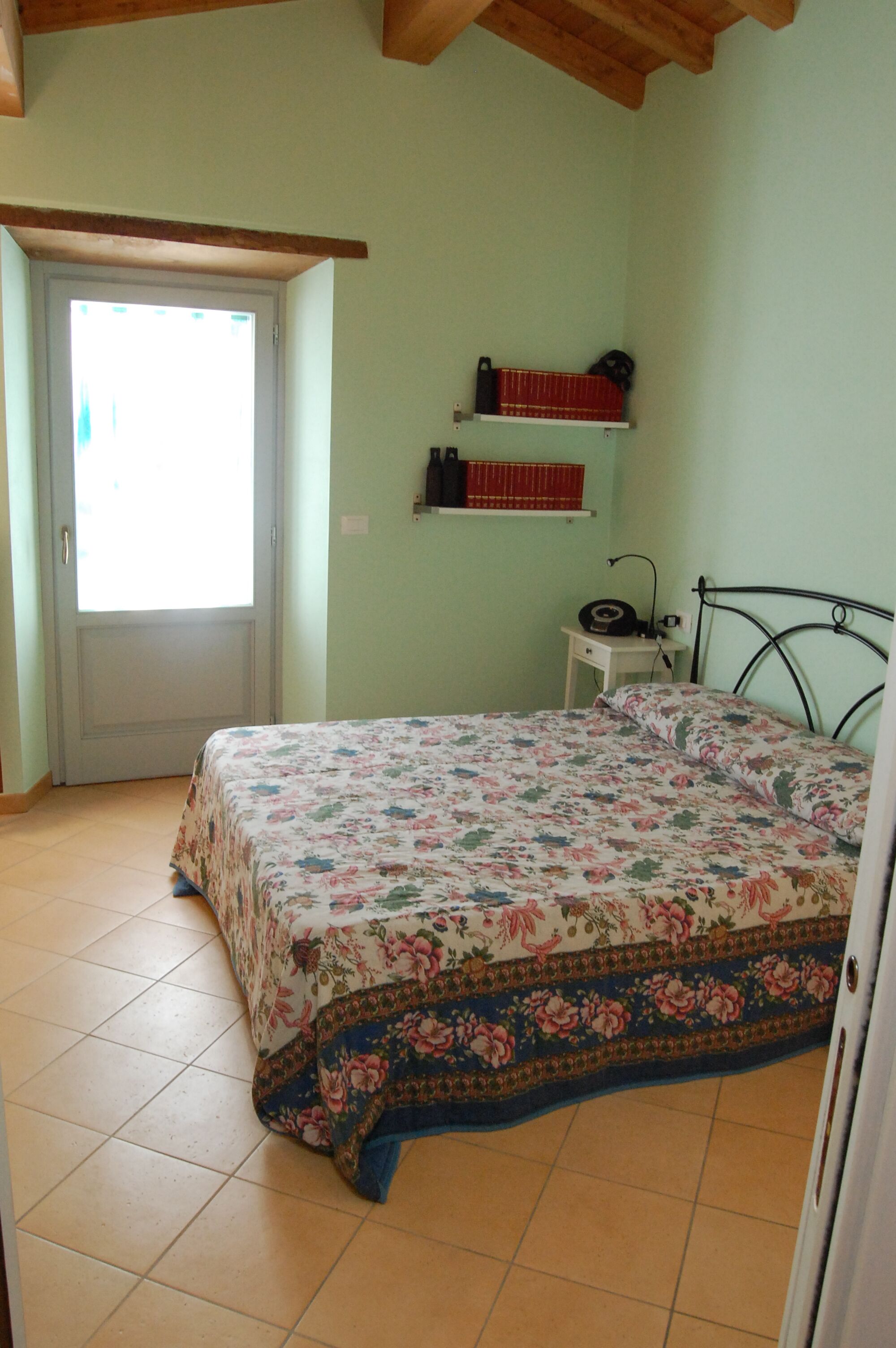 A bedroom with a bed, a door, a shelf with books, and a small table with a lamp.