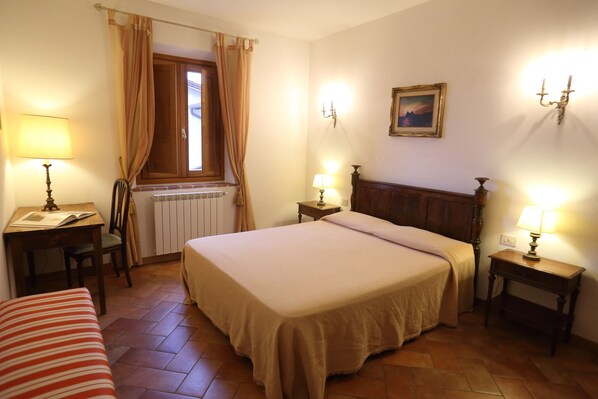 Un dormitorio con una cama, dos mesitas de noche con lámparas, una silla, una ventana con cortinas y un radiador.