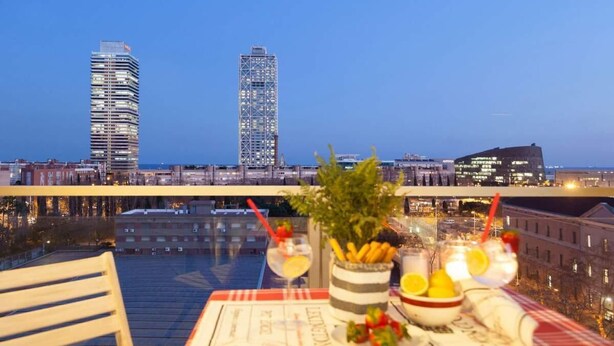 Un espace de restauration aménagé sur le toit, avec une table dressée pour deux personnes, offrant une vue sur un paysage urbain composé d’immeubles modernes et d’un plan d’eau.