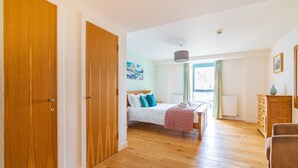 A bedroom with a wooden floor, a bed with white bedding and blue pillows, a dresser, and a window with curtains.