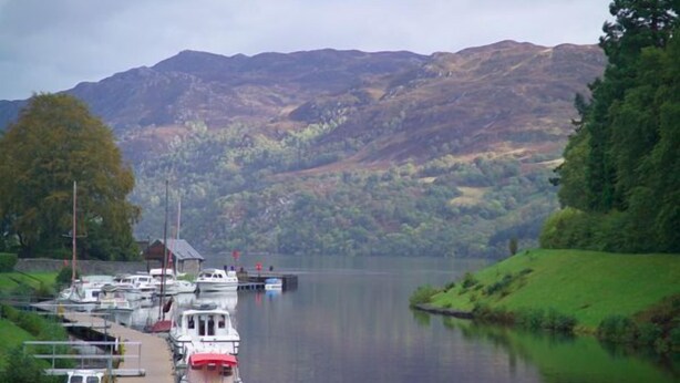 A calm lake with boats docked at a pier, surrounded by lush green hills.