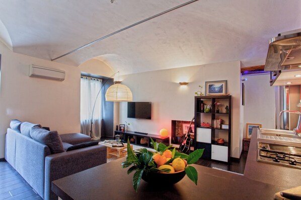A modern living room with a sofa, a dining table with a fruit bowl, a fireplace, and a bookshelf.