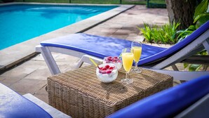 A poolside setting with a wicker table holding a bowl of fruit and two glasses of juice, a blue lounge chair, and a clear view of the pool.