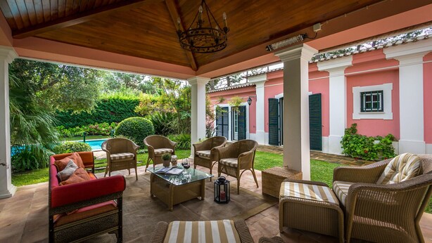A covered outdoor seating area with wicker furniture, a glass-top table, and a view of a swimming pool and greenery.