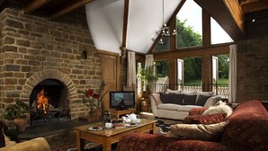 Un salon chaleureux avec une cheminée en pierre, des sièges confortables et de grandes fenêtres donnant sur l’extérieur.