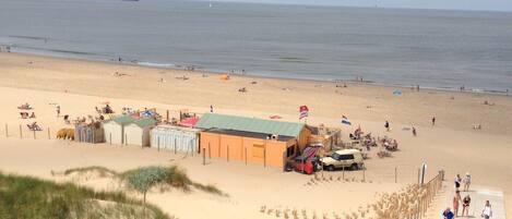 Une plage avec une promenade en bois menant à une zone sablonne où se trouvent des cabanes de plage et un drapeau.
