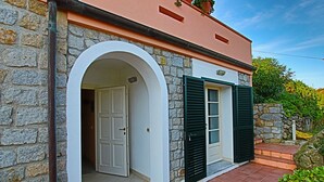 Un edificio de piedra con una puerta blanca y contraventanas verdes, un patio con baldosas y un cielo despejado.
